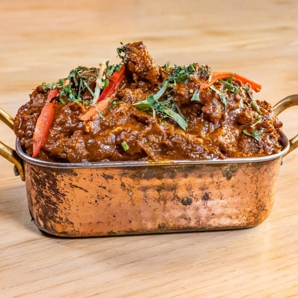 Rich, dark brown curry with red pepper strips and green herbs in a copper serving dish.