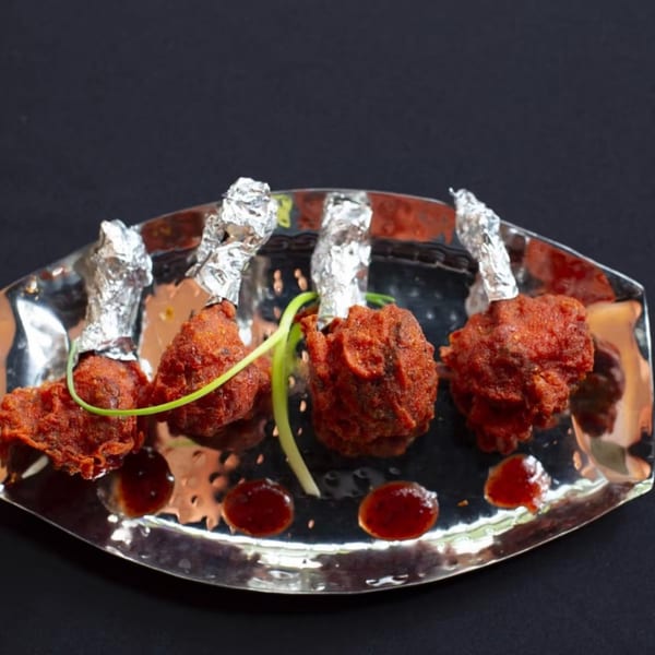 Four chicken lollipops with foil-wrapped bones, green garnish, and sauce on a silver platter.
