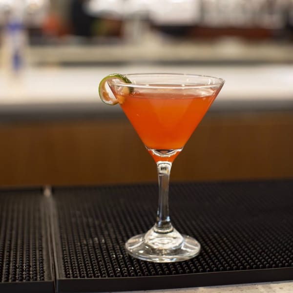 A vibrant red cocktail in a martini glass, garnished with a lime twist, on a bar mat.
