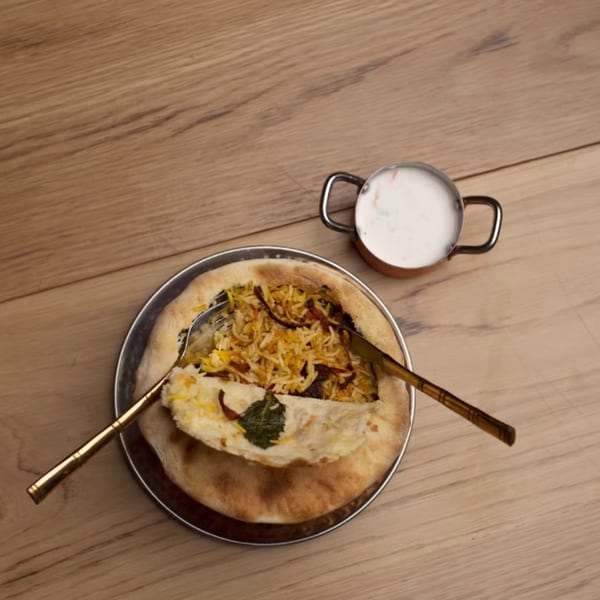 Biryani served in a bread bowl with a side of raita, on a wooden table.