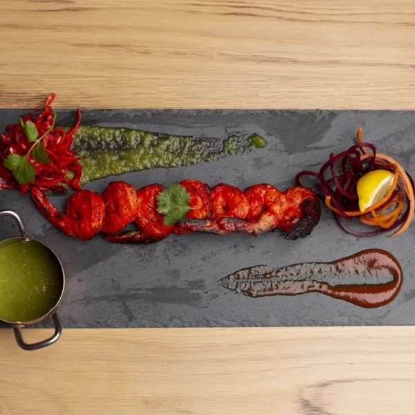 Grilled red shrimp with green sauce, red garnish, and lemon on a dark slate.