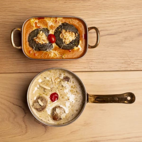 Two Indian dishes on a wooden table: a red curry with black dumplings and a creamy dessert with cherries.