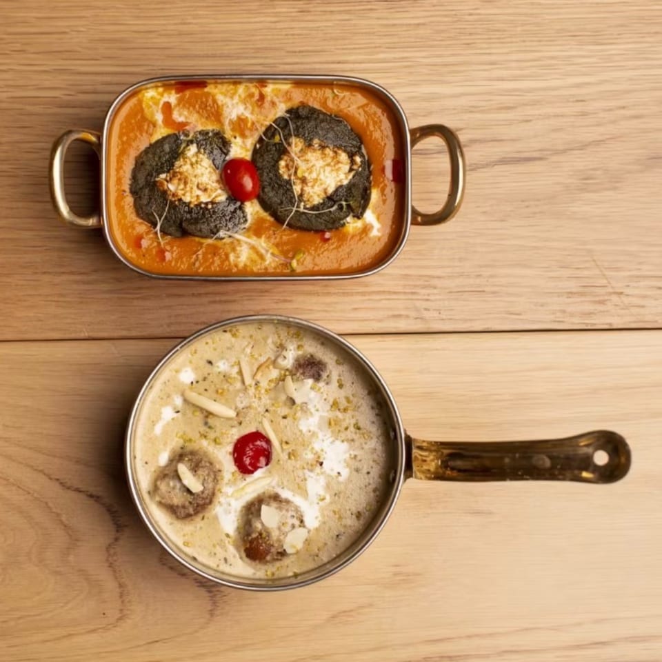 Two Indian dishes on a wooden table: a red curry with black dumplings and a creamy dessert with cherries.