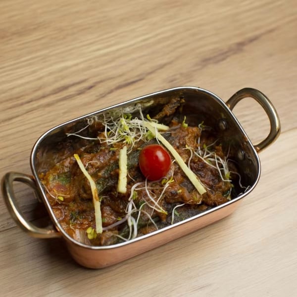 A copper-bottomed serving dish filled with a dark stew, garnished with sprouts, ginger, and a cherry tomato.