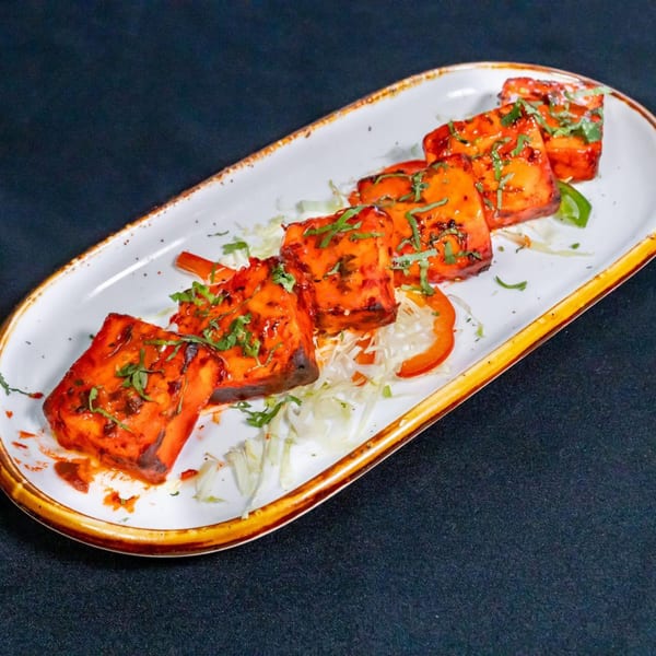 Six pieces of paneer tikka, garnished with fresh herbs and bell peppers, on a white oval plate.