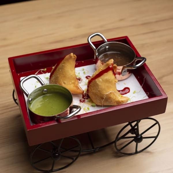 Two samosas with green and brown chutneys served in mini pots on a red cart.