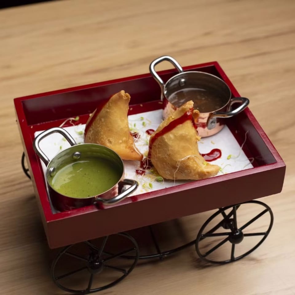 Two samosas with green and brown chutneys served in mini pots on a red cart.
