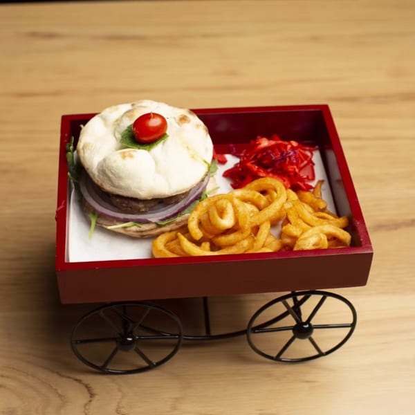 A burger with curly fries and red slaw served in a red wagon on a wooden table.